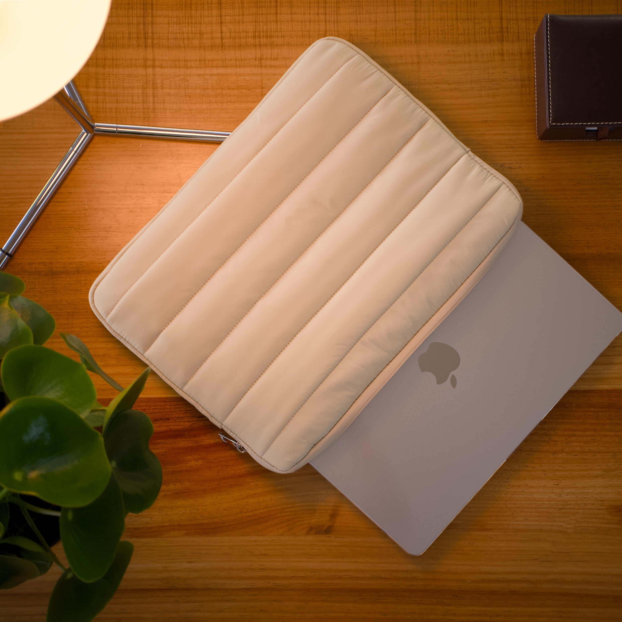 Beige padded laptop sleeve partially covering a MacBook on a wooden desk with a plant, lamp, and brown case nearby.
