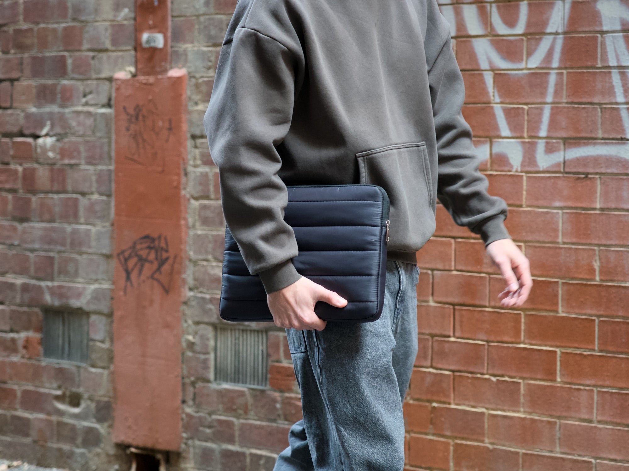 Man in grey hoodie holding a black Humley padded sleeve while walking through a brick laneway.