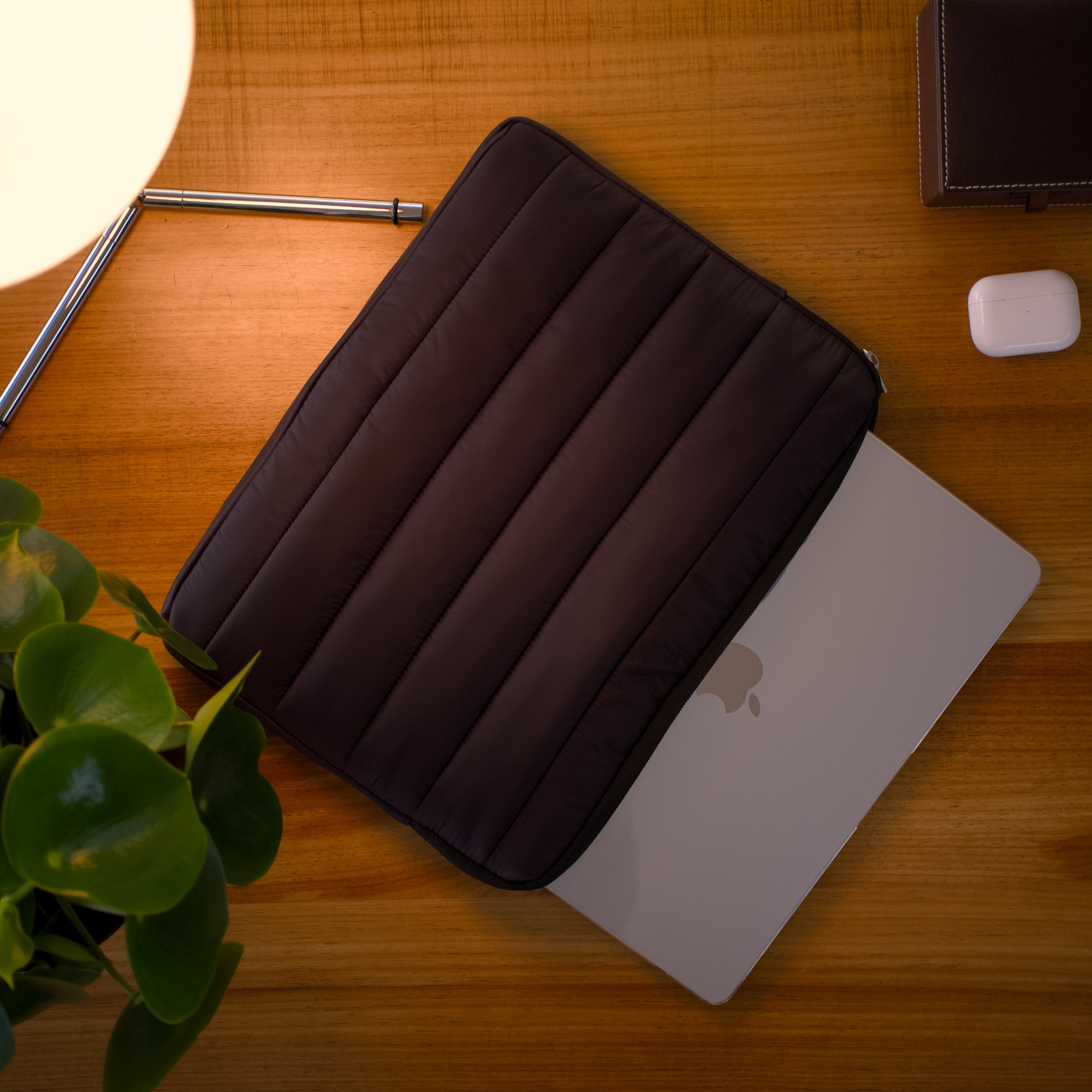 Black padded laptop sleeve partially covering a MacBook on a wooden desk with a plant, lamp, and AirPods nearby.