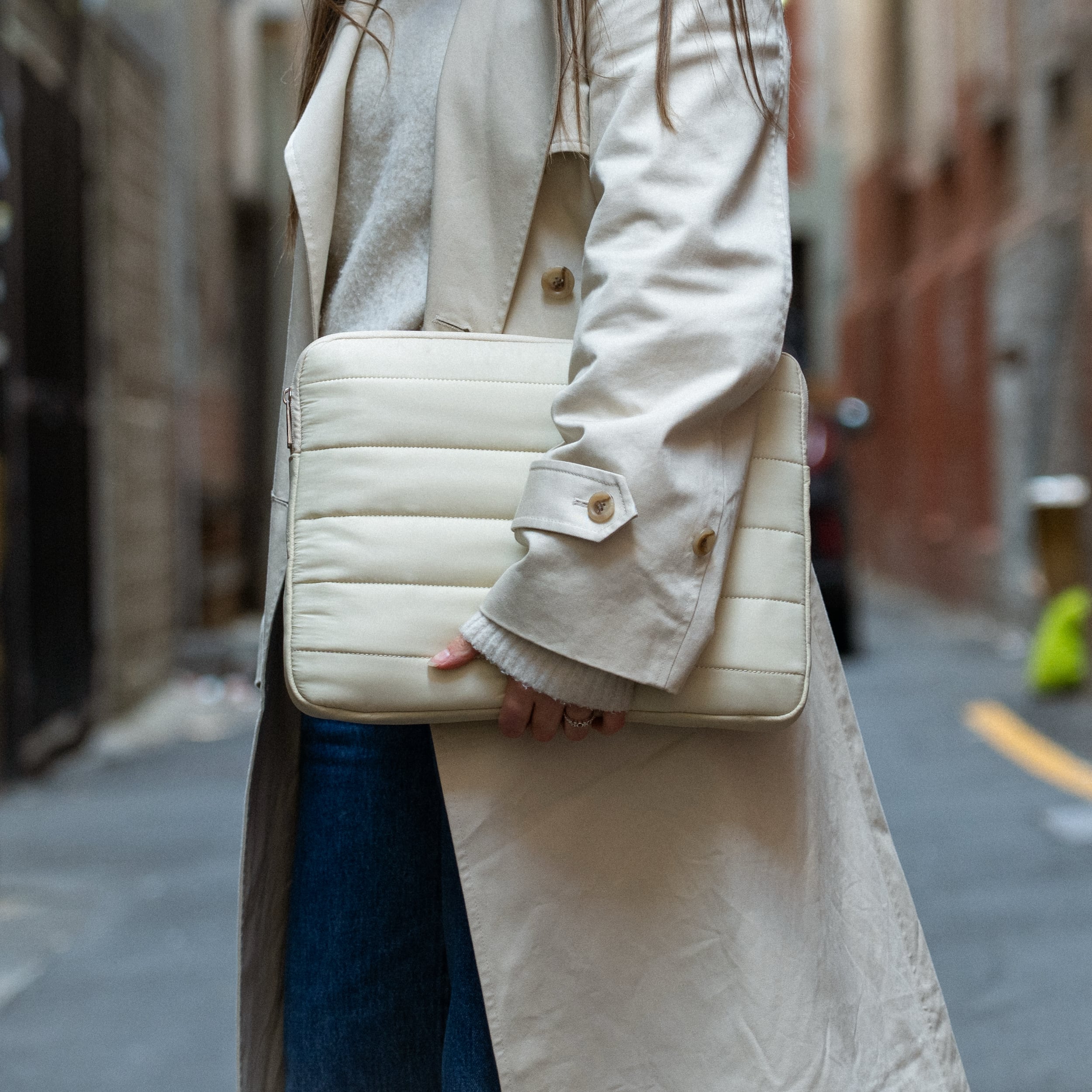 Woman holding a beige Humley padded laptop sleeve while walking through an urban alley in neutral outfit