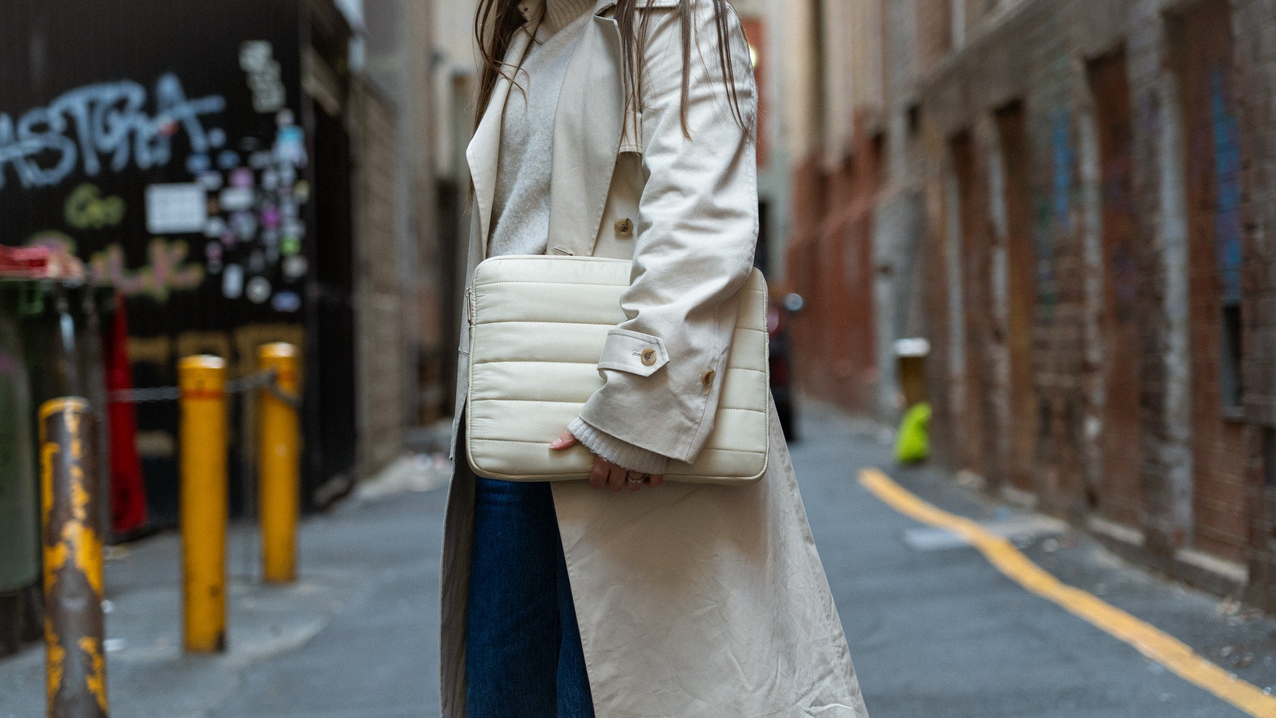 Woman holding a beige Humley padded laptop sleeve while walking through a graffiti-lined urban alley
