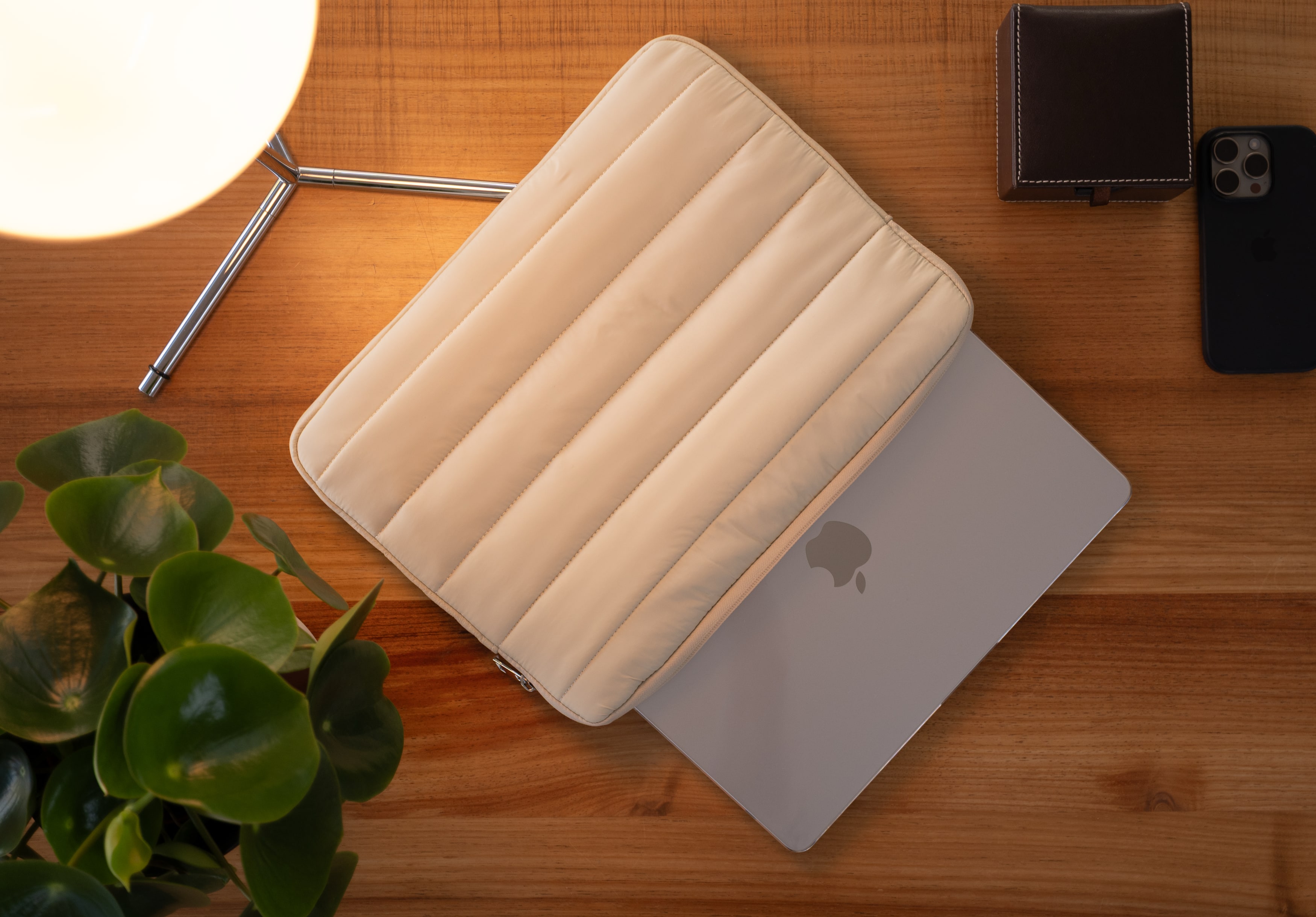 Top-down view of a beige Humley padded sleeve partially covering a MacBook on a wooden desk with a plant and accessories.