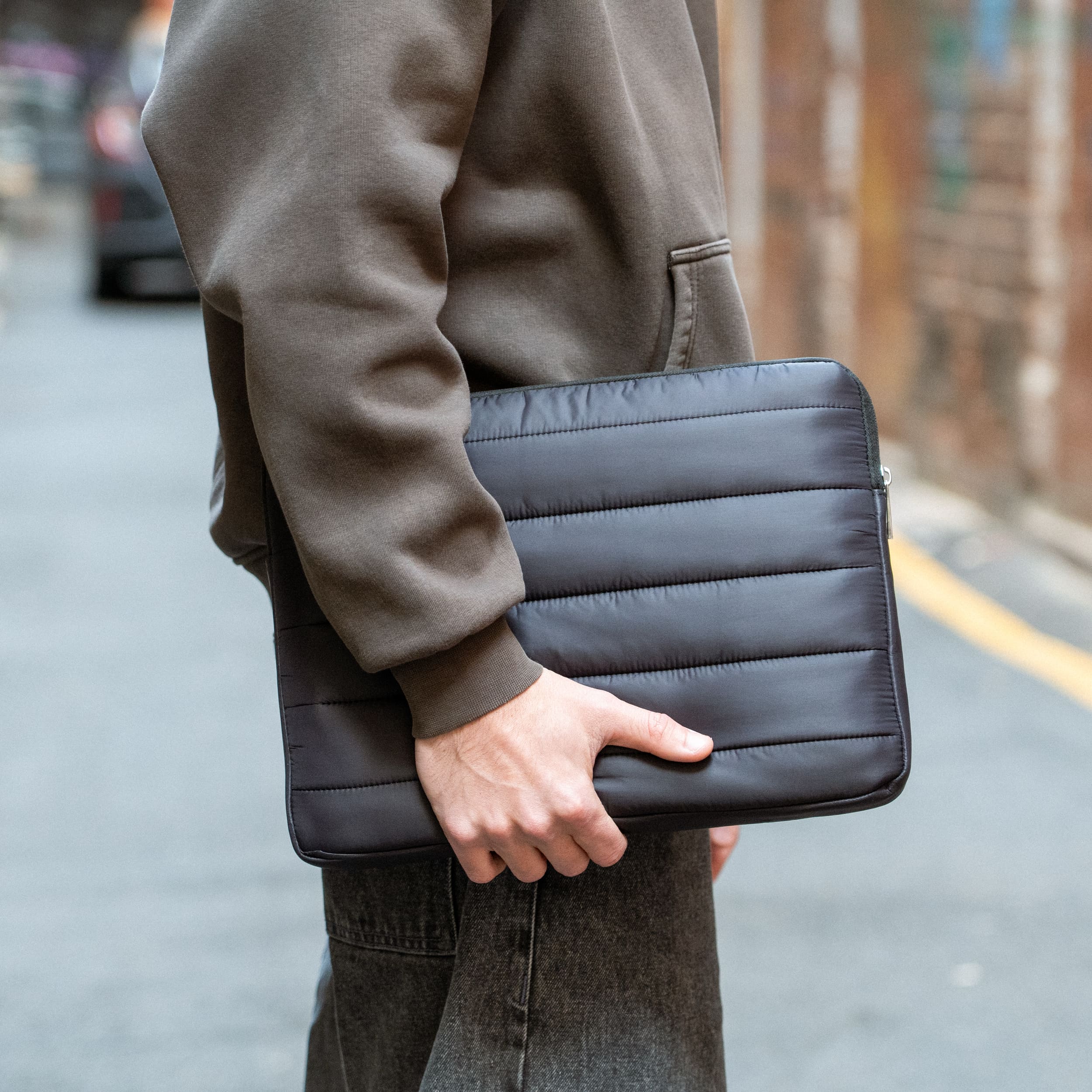 Man holding a black Humley padded laptop sleeve in urban alley setting with casual outfit
