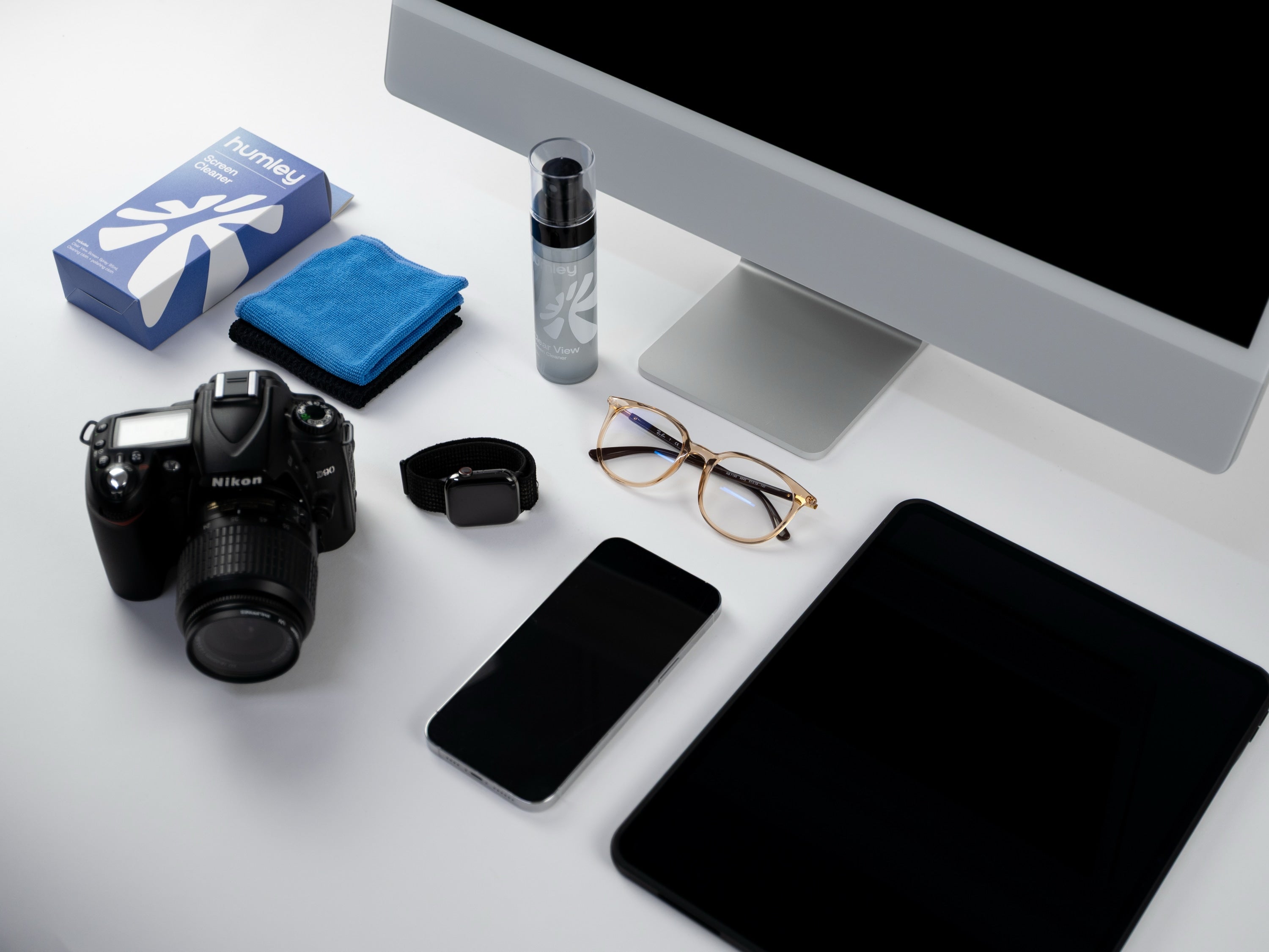 Flat lay of Humley screen cleaner kit with an iMac, camera, smartwatch, smartphone, tablet, eyeglasses, and microfibre cloths on a white desk.